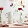 A white teapot and cups are set on a pink tablecloth, with flowers in the background. Produktbild-Vorschau