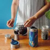 A person is shown scooping loose tea leaves from a jar into a small metal container, with a patterned thermal cup and another jar of tea in the foreground. Anteprima dell’immagine prodotto