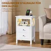 A white nightstand with drawers and an open shelf, featuring a phone charging on top and a flip clock in the shelf, placed next to a sofa. Produktbild-Vorschau