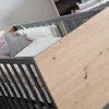 Close-up view of a crib with bedding and a wooden panel. Produktbild-Vorschau