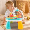A baby is playing with a colorful interactive music table. The baby is on its hands and knees, reaching for the toy. The background includes a knitted blanket and wooden blocks. Produktbild-Vorschau