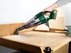 A person is using a cordless handheld vacuum cleaner to clean sawdust off a wooden workbench. Produktbild-Vorschau