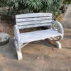 A garden bench with a rustic, wagon-wheel design is placed outdoors on a paved area, with trees and foliage in the background. Produktbild-Vorschau
