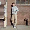 A young man stands leaning against a brick wall next to a metal shuttered door. Anteprima dell’immagine prodotto