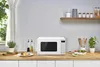 A white microwave oven is placed on a wooden countertop in a kitchen setting, with food and kitchenware around it. Produktbild-Vorschau