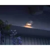 A motion-activated LED downlight is mounted on the underside of a roof overhang, illuminating the exterior of a house at night. Some purple flowers are visible in the foreground. Voir les images du produit
