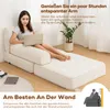 A woman is using a laptop while sitting on a modular sofa that can be converted into a bed. The sofa is placed near a wall, and there is a plant and a small table with a coffee cup and a tablet. Podgląd zdjęcia produktu