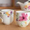 Two ceramic mugs with floral designs are placed on a wooden surface. A teapot is visible in the background. Podgląd zdjęcia produktu