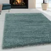 A shaggy rug in an aqua color is displayed in a living room setting with a fireplace and a blue ottoman. Produktbild-Vorschau
