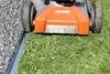 A close-up view of a lawnmower's front, with its wheels on a grassy area next to a gravel path separated by a metal edge. Produktbild-Vorschau