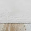 A close-up view of a white, high-pile rug with a scalloped edge, resting on a light brown wooden floor. Produktbild-Vorschau