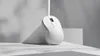 A white wireless mouse is shown on a white surface with shadows cast on a gray background. Produktbild-Vorschau