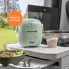 A mini rice cooker is shown on a table outdoors, with a bowl of food and other kitchen items. Produktbild-Vorschau