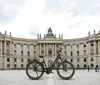 An e-bike is parked in front of a grand, classical building on a cobblestone plaza. Produktbild-Vorschau