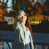 A woman is taking a selfie with her phone, which has a lanyard attached, in an outdoor park setting with autumn foliage. Anteprima dell’immagine prodotto