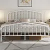 A white metal bed frame with a crown-shaped headboard, set up in a bedroom with pillows and a rug. Produktbild-Vorschau