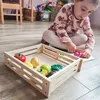 A young girl is playing with a wooden toy crate filled with toy fruits and vegetables. Náhled obrázku produktu