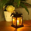 A solar-powered lantern with a candle inside sits on a wooden table next to a potted plant with a yellow rose. Voir les images du produit