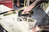 A person is using a cordless jigsaw to cut a curved line on a wooden board in a workshop setting. Tools like a hammer and pencils are visible on the board. Podgląd zdjęcia produktu