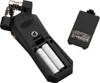 Close-up view of a black audio recorder with its battery compartment open, showing two AA batteries inserted. The battery cover is detached and placed to the side. Produktbild-Vorschau