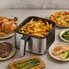 An air fryer filled with french fries is surrounded by various cooked foods, including a whole chicken, steak, salmon, asparagus, broccoli, and pizza. Náhled obrázku produktu