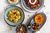 A flat lay of various dishes, including roasted potatoes, dates with cheese, roasted pumpkin, chestnuts, and mushrooms, arranged on a light-colored surface. Produktbild-Vorschau