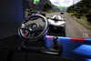 A racing game setup with a steering wheel and joystick in the foreground, and a racing game displayed on a large screen in the background. Anteprima dell’immagine prodotto