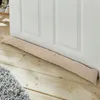 A beige fabric draft stopper lies on a wooden floor in front of a white door. Produktbild-Vorschau