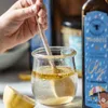 A hand is stirring a mixture in a glass jar with a wooden spoon. A slice of lemon is in the foreground, and a bottle of oil is to the right. Produktbild-Vorschau
