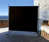 A black privacy screen stands on a paved patio with a stone wall and the ocean in the background. Produktbild-Vorschau