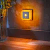 A night light is plugged into a wall outlet, casting a warm amber glow on the wall and floor. A vase with yellow flowers is visible to the left. Produktbild-Vorschau