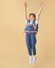 A young girl is jumping in the air with her arms raised, wearing a backpack and overalls. Anteprima dell’immagine prodotto
