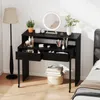 A black vanity desk with drawers and makeup products is shown in a bedroom setting next to a bed and a floor lamp. Náhled obrázku produktu