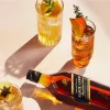 A bottle of Johnnie Walker Black Label whisky and two cocktails with garnishes, arranged on a pink surface with dramatic shadows. Produktbild-Vorschau
