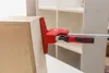 A woodworking clamp is shown holding together two pieces of wood, likely part of a cabinet or furniture construction. In the background, partially visible, are unfinished shelving units. Produktbild-Vorschau
