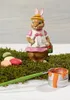 A ceramic Easter bunny figurine dressed in a hat and apron, holding a paintbrush and a bucket of paint, surrounded by moss and colorful Easter eggs. Podgląd zdjęcia produktu