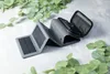 A gray foldable solar charger with a power bank and wireless charging capability is laid out on a white surface, surrounded by green eucalyptus leaves. Produktbild-Vorschau