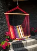 A colorful striped hanging chair is suspended from a hook, set against a dark stone wall. It is placed on a set of outdoor steps, with potted flowers on either side. Náhled obrázku produktu