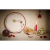 A festive Christmas display on a shelf, featuring a large decorative plate, a rocking horse, gingerbread cookies, and candles. Náhled obrázku produktu