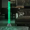 A green LED water column lamp with decorative fish is placed in a modern living room next to a leather sofa. Produktbild-Vorschau