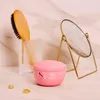 A pink hair mask jar is displayed on a marble surface, alongside a wooden hairbrush in a glass holder and a gold vanity mirror. Voir les images du produit