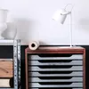 A white desk lamp is placed on a wooden cabinet next to a metal shelf with vases and a roll of paper. Produktbild-Vorschau