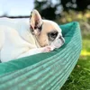 A French Bulldog is resting in a green corduroy dog bed outdoors on the grass. Náhled obrázku produktu