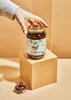 A hand reaching for a jar of olives placed on a cube, with a few olives in a small dish in the foreground. Produktbild-Vorschau