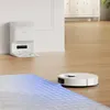 A white robot vacuum and its charging dock are shown on a wooden floor, with a blue grid overlay indicating cleaning coverage on a rug. Produktbild-Vorschau