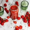 A glass jar filled with tomato sauce is surrounded by fresh cherry tomatoes and peppercorns on a white wooden surface, with two green glasses in the background. Produktbild-Vorschau
