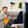 A man is sitting on a couch, holding a remote control and a mug, while watching TV. Voir les images du produit