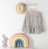 A child's striped shirt with a peplum hem, a straw hat, and a rainbow pillow are displayed against a white wall. Produktbild-Vorschau