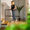 A young woman is carrying a foldable drying rack down a hallway with stairs on the left and a door on the right. Náhľad obrázku produktu