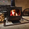 A black cast iron wood-burning stove with a fire burning inside, placed on a wooden floor. To the left, a basket filled with firewood. In the background, two chairs and a wall. Anteprima dell’immagine prodotto